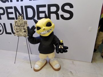 A yellow and black mascot figure in a hoodie stands next to a cardboard cutout of itself at what appears to be a convention booth with 'SPENDERS' visible in the background.