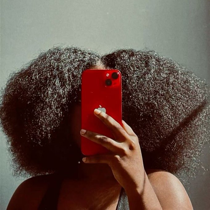 A person with voluminous, curly natural hair takes a mirror selfie, obscuring their face with a red iPhone.