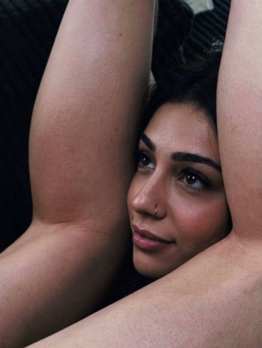 A close-up portrait of a woman with dark hair, a nose ring, and glossy lips, resting with her arms raised above her head.