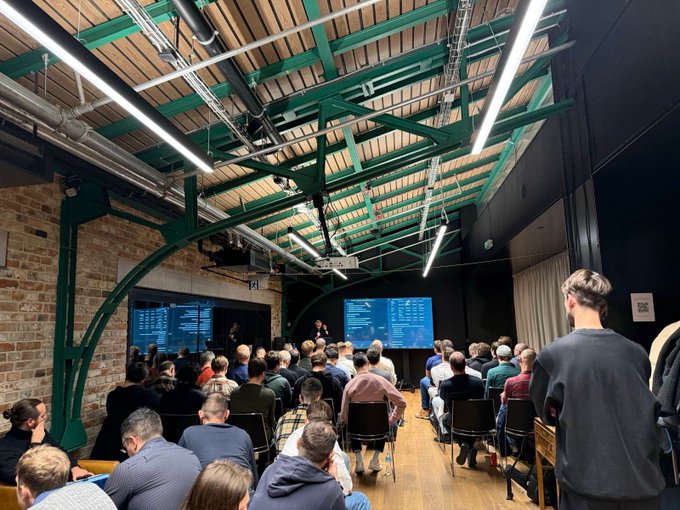 A presentation or meetup event in an industrial-style venue with exposed brick walls, green metal beams, and hanging tube lights, where an audience sits facing two presentation screens displaying data.