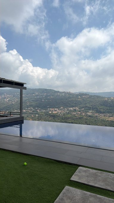 A photograph of an infinity pool overlooking a hillside town with a tennis ball on artificial grass in the foreground.