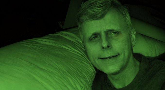 A selfie taken at night with a strong green night-vision tint, showing a man with short light hair lying against a white pillow, smiling softly at the camera.