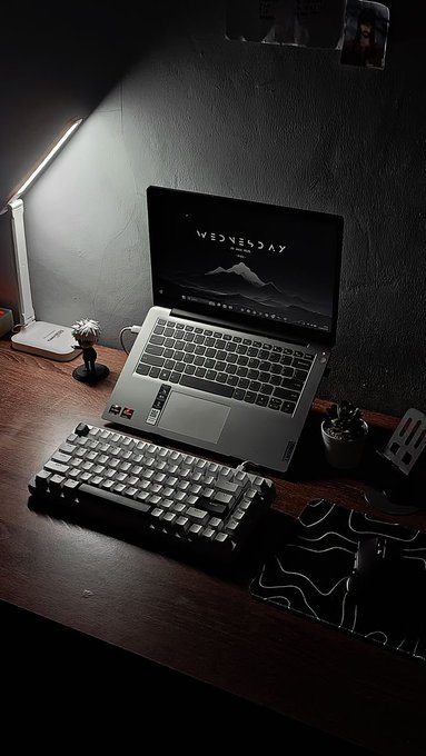 A moody, dimly lit desk setup featuring an open laptop displaying the Wednesday wallpaper, an external keyboard, a small figurine, and a plant illuminated by a desk lamp.