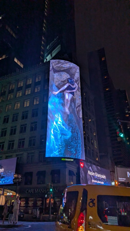 A nighttime city street view featuring a large vertical LED billboard displaying an artistic image of a figure with blue flowing water, with a yellow taxi and "CAFE MANHA" storefront visible below.