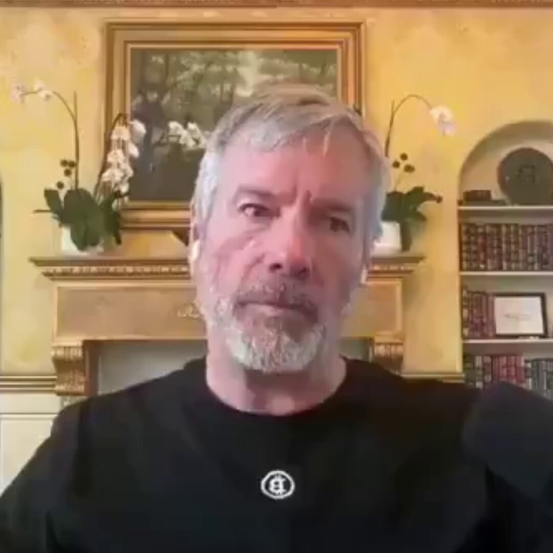 The image features a man wearing a black shirt who appears to be giving a speech or interview, with his face clearly visible against the backdrop of a room filled with bookshelves. The room has an elegant atmosphere, as evidenced by the presence of potted plants and other decorative elements such as vases placed on the shelves. The image also contains text that reads "YOU NEED TO SEE THIS MICROSTATEGY WILL EVENTUALLYOWN 5% OF THE entire Bitcoin supply.". This suggests that the man's speech or interview might be related to a topic involving cryptocurrency, specifically Bitcoin, and its potential impact on the global economy.