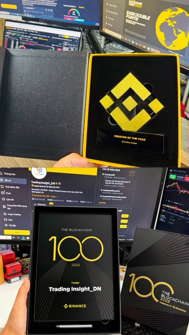 The image features a person holding up two items - an award and a book titled "The 100 Million Views" by Binance Square. The person is also holding a computer monitor displaying trading insights, which is part of their work at Binance Square. The background shows a desk with various screens and keyboards, indicating that the person might be involved in the world of cryptocurrency or financial technology.