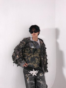 The image features a person wearing a camouflage jacket, sunglasses, and holding a bag with a white star design. The person's face is partially visible behind them as they pose for the photo. The background of the image consists of a plain white wall, which contrasts with the colorful elements in the foreground. The post that includes this image has been edited to include a watermark that reads "x = #ALPHADRIVEONE #ALD1 #알파드라이브원 프리에서 작성자로 변환".