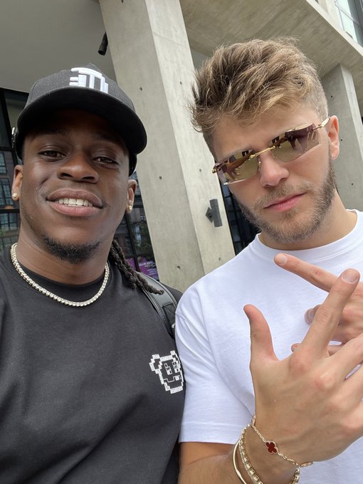 Two men pose together outdoors—one wearing a black cap with a logo and graphic tee smiling broadly, the other in a white t-shirt and yellow-tinted sunglasses throwing up a hand gesture—aligning with the post's promotion of a live crypto bull market alpha call.