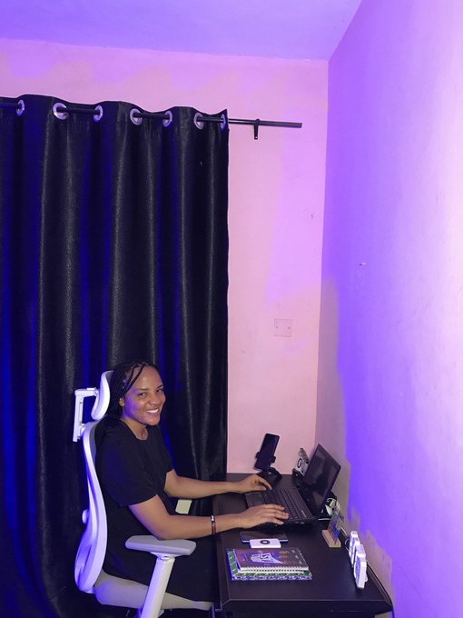 A smiling woman sits at a desk in a room illuminated with purple and pink lighting, working on a laptop next to dark curtains.