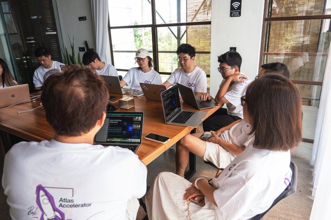 A group of young people wearing matching white t-shirts sit around a wooden table working on their laptops during what appears to be a collaborative session. The back of one shirt reads 'Atlas Accelerator', indicating this is likely a startup or entrepreneurial program.