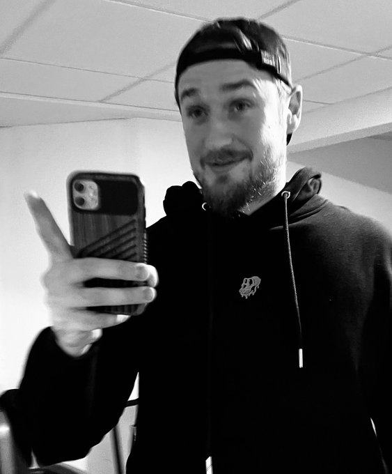 A black-and-white selfie of a bearded man wearing a backward baseball cap and a dark hoodie, holding up his smartphone and pointing at it.