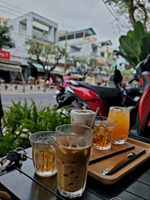 An inviting café scene in Vietnam features four refreshing drinks—an iced coffee, a creamy espresso beverage, iced tea, and orange juice—arranged on a wooden tray beside a set of keys, with motorbikes and bustling street life visible in the background. The relaxed "Gm" greeting pairs perfectly with this casual morning setup, suggesting a leisurely coffee break to start the day.