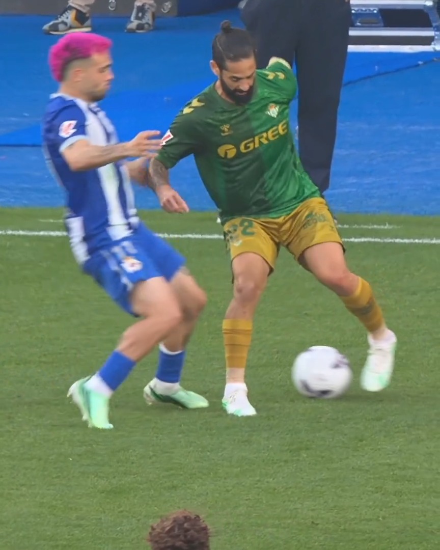 Two soccer players compete for the ball during a match; one wears a blue and white striped kit with bright pink hair, while the other wears a green jersey with 'GREE' sponsor text and yellow shorts with the number 22.