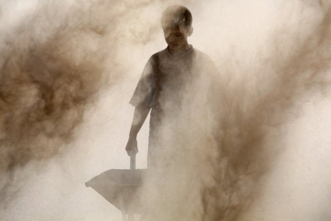 A man pushing a wheelbarrow is obscured by a large, thick cloud of brown dust or smoke.