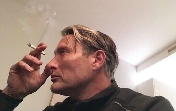 A selfie-style photo of Mads Mikkelsen smoking a cigarette indoors.