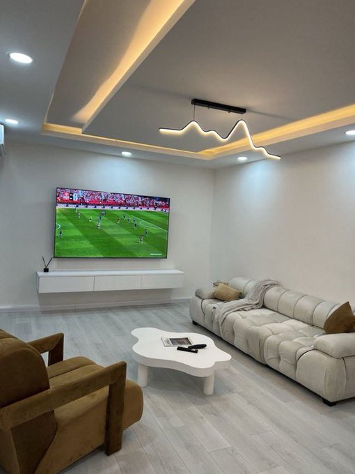 The image shows a living room with white walls and hardwood floors. A flat screen TV is mounted on the wall, displaying a soccer game. There are two couches in the room - one large couch occupying most of the space and another smaller couch placed against the opposite wall. The room also features a coffee table near the larger couch, as well as a chair positioned next to it.
