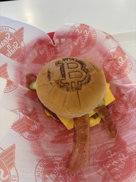 The image features a burger with bacon, cheese, and lettuce inside of a bun. The burger has been adorned with a Bitcoin symbol or logo, which adds a unique touch to the meal. The burger is placed on top of a red paper that displays the words "Had the bitcoin burger @SteaknShake" and "Had a lot of trouble with my @AquaBitcoin @Excellion wallet - which has happened many times at many bitcoin merchants". This suggests that someone might have used Bitcoin to purchase this meal, possibly as part of an online transaction or a Bitcoin-themed restaurant. The image also includes the words "Had the bitcoin burger @SteaknShake" and "Had a lot of trouble with my @AquaBitcoin @Excellion wallet - which has happened many times at many bitcoin merchants", indicating that this is likely a humorous or satirical post related to Bitcoin, food, or both.
