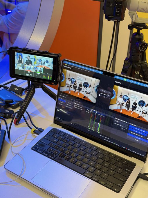 A laptop displays live streaming software showing a panel discussion, with a monitor mounted on a tripod beside it and professional video equipment in the background.