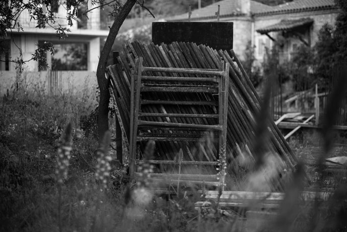 A black and white photograph of a makeshift wooden structure or ramp standing in overgrown grass, with residential buildings visible in the background.