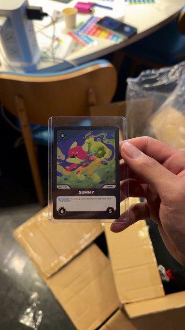 A hand holds a Pokemon card featuring GUMMY in a protective sleeve, showing 6 HP and 5 damage, set against a blurred office background with cardboard boxes.