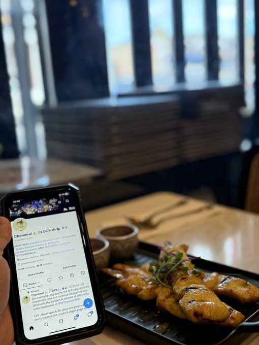 A hand holds a smartphone displaying the Clock In BBL Instagram profile at 'Chanimal,' positioned next to a plated meal of chicken wings in what appears to be a restaurant setting.