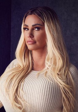 A portrait of a blonde woman with long wavy hair wearing a cream-colored knit sweater, posing against a dark background.
