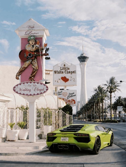 Street scene featuring a bright green Lamborghini parked in front of the 'Little White Chapel' and 'A Little White Wedding Chapel' signs in Las Vegas, with the Stratosphere tower visible in the background.