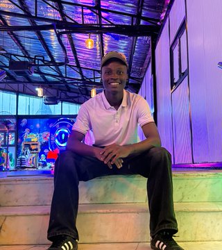 A photograph of a smiling young man wearing a pink polo shirt and a cap, sitting casually on steps in what appears to be an arcade or entertainment venue with neon lighting and game machines in the background.
