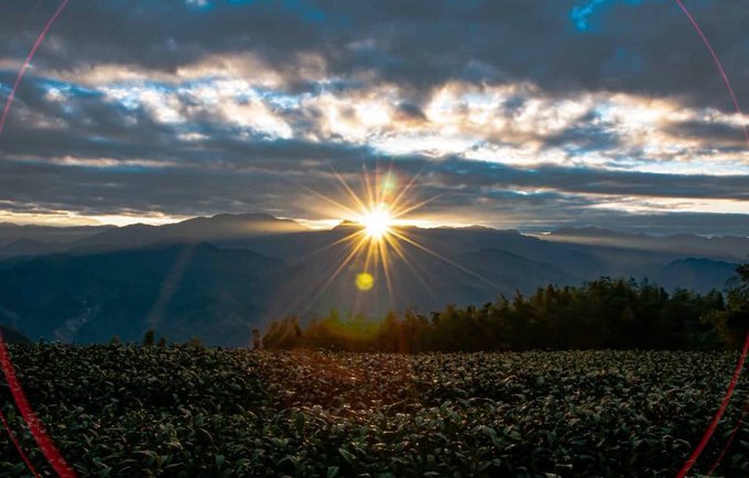 A scenic landscape photograph captures a bright sun setting behind a cloudy sky, casting rays over distant mountains and a foreground field.