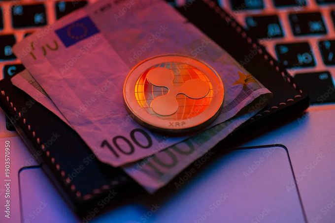 A close-up photo of a physical XRP (Ripple) cryptocurrency coin resting on a 100 Euro banknote on a black wallet placed on a laptop keyboard, with red and blue lighting.