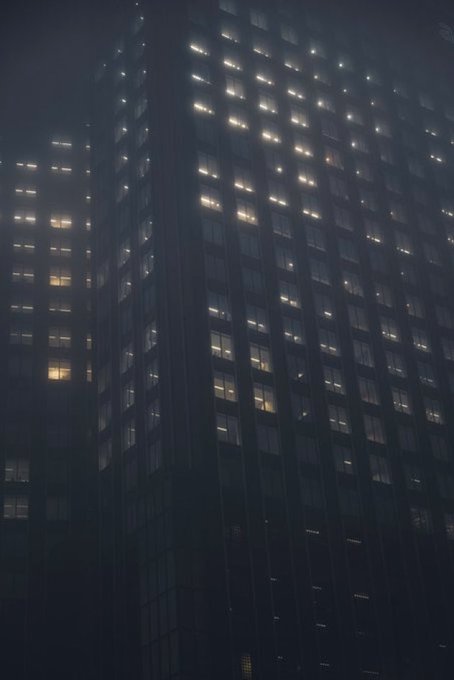 A nighttime view of a tall skyscraper with a grid pattern of illuminated windows glowing against the dark facade. The building appears to be an office tower with scattered lights still on, suggesting late evening or after-hours activity.