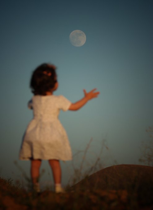 A softly blurred photograph of a small child in a white dress standing on a hill at dusk, reaching her hand up toward a full moon in the twilight sky.