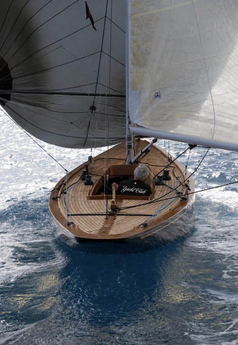 A classic wooden sailboat named "Giudecca" glides through open water, captured from an aerial perspective showing its polished deck, expansive white sails, and the deep blue sea surrounding it.