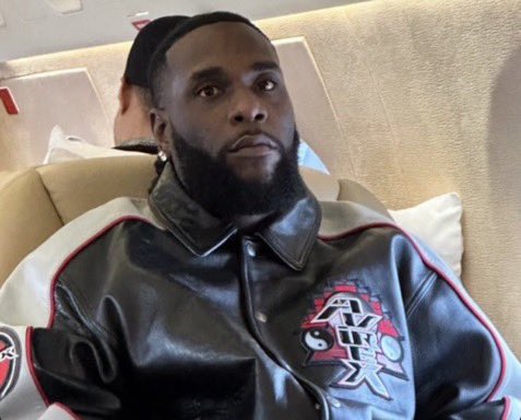 A photograph of a man with a beard wearing a black beanie and a black leather jacket with Japanese characters, sitting in what appears to be an airplane cabin.