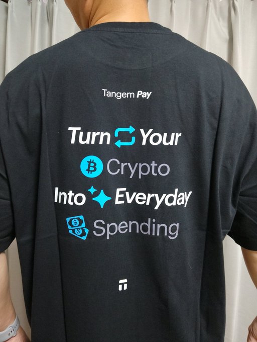 The image shows a person wearing a black t-shirt with white text that reads "turn your daily spending into tangem". It appears that this individual might be promoting the concept of turning everyday expenses, such as shopping or dining out, into virtual currency through an app like tangem. This can be seen as a way to manage spending more efficiently and potentially earn some extra money in return for participating in these activities on the app.