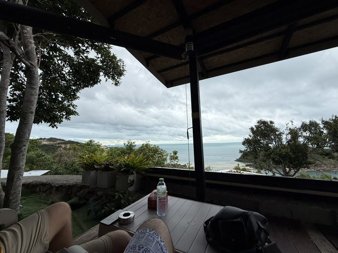 A scenic view from a covered patio overlooking a calm ocean, with a person's legs visible, a wooden table holding a water bottle and ashtray, and a bag nearby.