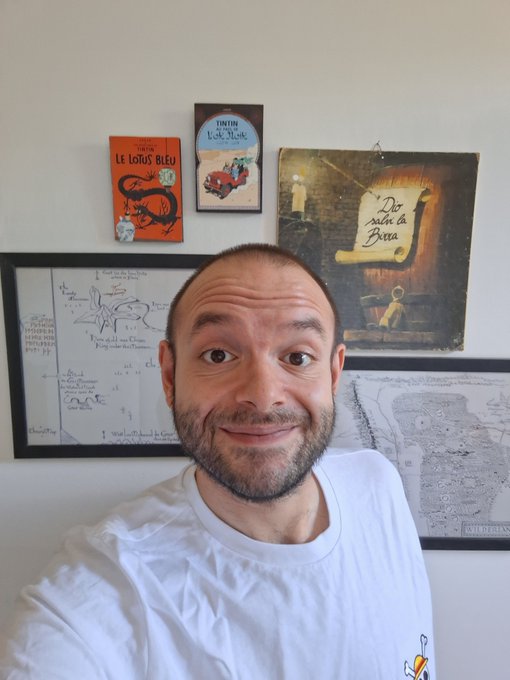 A man taking a selfie in front of a wall decorated with children's books in French including 'Le Loup Bleu' and 'Un Petit Rêve', alongside framed artwork and sketches.