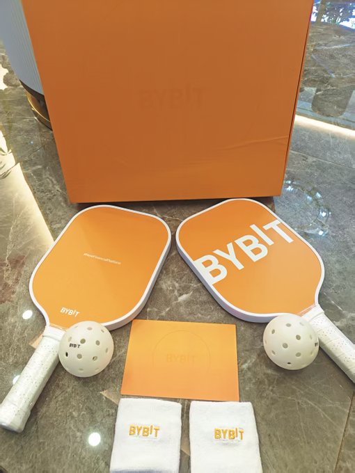 BYBIT branded pickleball paddles and balls with an orange gift bag and BYBIT branded wristbands displayed on a marble surface.