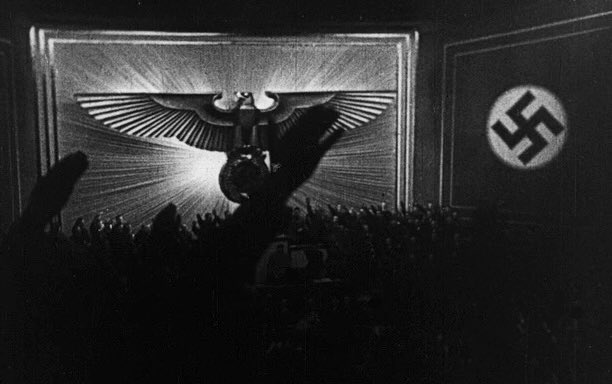 A crowd of people raise their arms in a Nazi salute during a rally, beneath a large eagle emblem and a swastika flag. The image appears to be from a propaganda film or photograph from Nazi Germany.