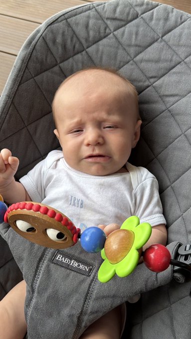 An adorable baby with a tired, fussy expression sits in a gray "BAD BJORN" baby bouncer, clutching a colorful wooden toy with red, green, and blue pieces across their lap. The baby appears overwhelmed and ready for a meltdown, perfectly matching the desperate cry for help from a sleep-deprived parent seeking advice on how to soothe their cranky little one.