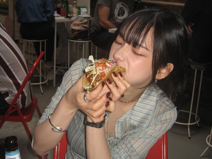 A young woman with dark hair and bangs enthusiastically bites into a hot dog topped with sauce and cabbage while sitting at a restaurant table.