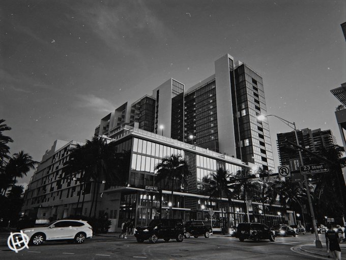 A black and white nighttime cityscape showing a modern multi-story building at an intersection marked '23 Street' with a 'No Turn' sign, illuminated streetlights, and parked cars.