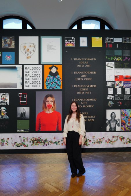 A woman poses in an art gallery in front of a black wall display featuring text that reads 'I TRANSFORMED IDEAS INTO ART / I TRANSFORMED ART INTO CODE / I TRANSFORMED CODE INTO NFT / I TRANSFORMED NFT TO MONEY.'
