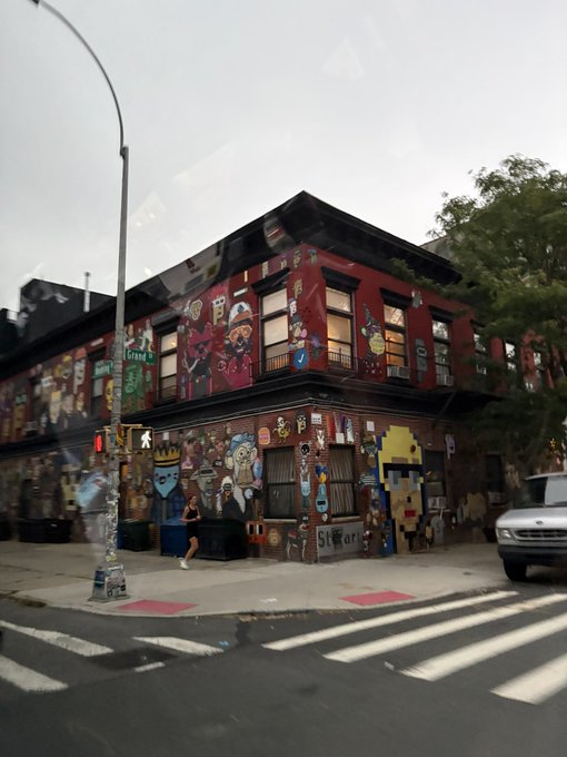 A corner building in Williamsburg is covered in colorful street art and character murals, with visible text reading "GRAND" and "Sklar" on the graffiti-covered façade. The image illustrates the post's point perfectly—despite constant turnover in the neighborhood, this long-standing NFT (not-for-tourism) tagged building remains a fixture in 2026.