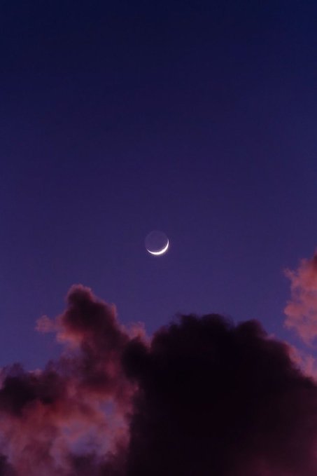 A crescent moon glows in a deep purple twilight sky above dark, pink-tinged clouds.