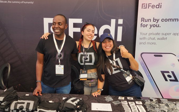 Three smiling Fedi team members pose together at a promotional booth, with the woman on the right giving a thumbs-up in front of a "Fedi" banner advertising the app as "your private super app with chat, wallet, and more." The image captures the LATAM-based team after nearly three and a half years of building Fedi, with branded merchandise visible on the table in front of them.