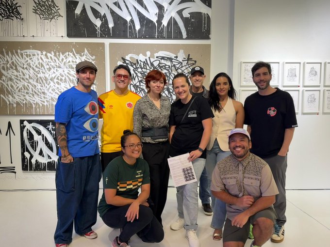 A group of nine people pose together in a gallery space, standing and kneeling in front of wall-mounted black-and-white graffiti-style artworks on the left and framed drawings on the right.