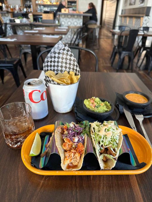 The image shows a table with three tacos, two of which are placed on a yellow plate while one is on a white plate. There is also a glass of beer next to the plates. The table appears to be located inside a restaurant or bar setting, as there are chairs and dining tables visible in the background.