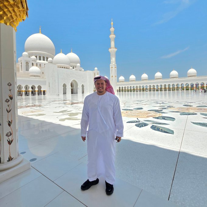 A man in traditional Emirati attire (white kandura and red-checkered ghutra) stands smiling in the courtyard of the Sheikh Zayed Grand Mosque, with its iconic white domes, minarets, and intricate floral marble inlay visible behind him. The image's grandeur and symbolism of visionary achievement mirror the post's themes of ambition, legacy, and redefining boundaries—whether in architecture, finance, or human potential.