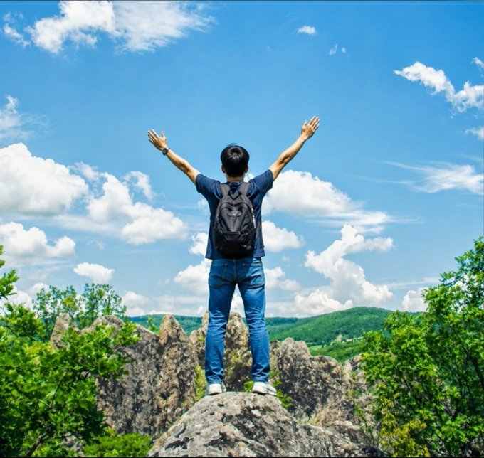 A person wearing a backpack stands triumphantly on a rocky outcrop with arms raised toward a bright, cloudy sky, surrounded by green hills.